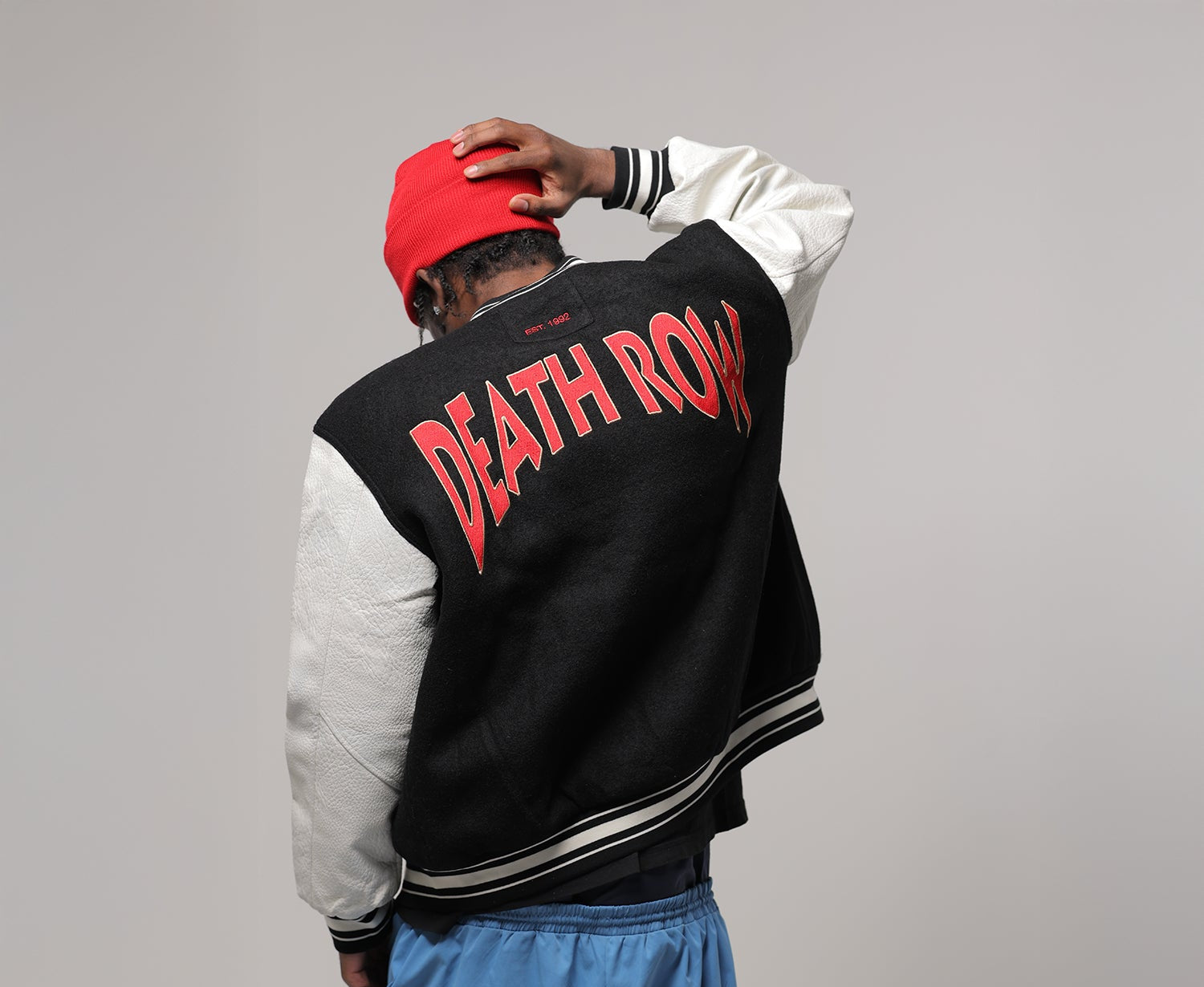 Person wearing a black and white varsity jacket with 'DEATH ROW' text on the back, red cap, and blue pants against a gray background.