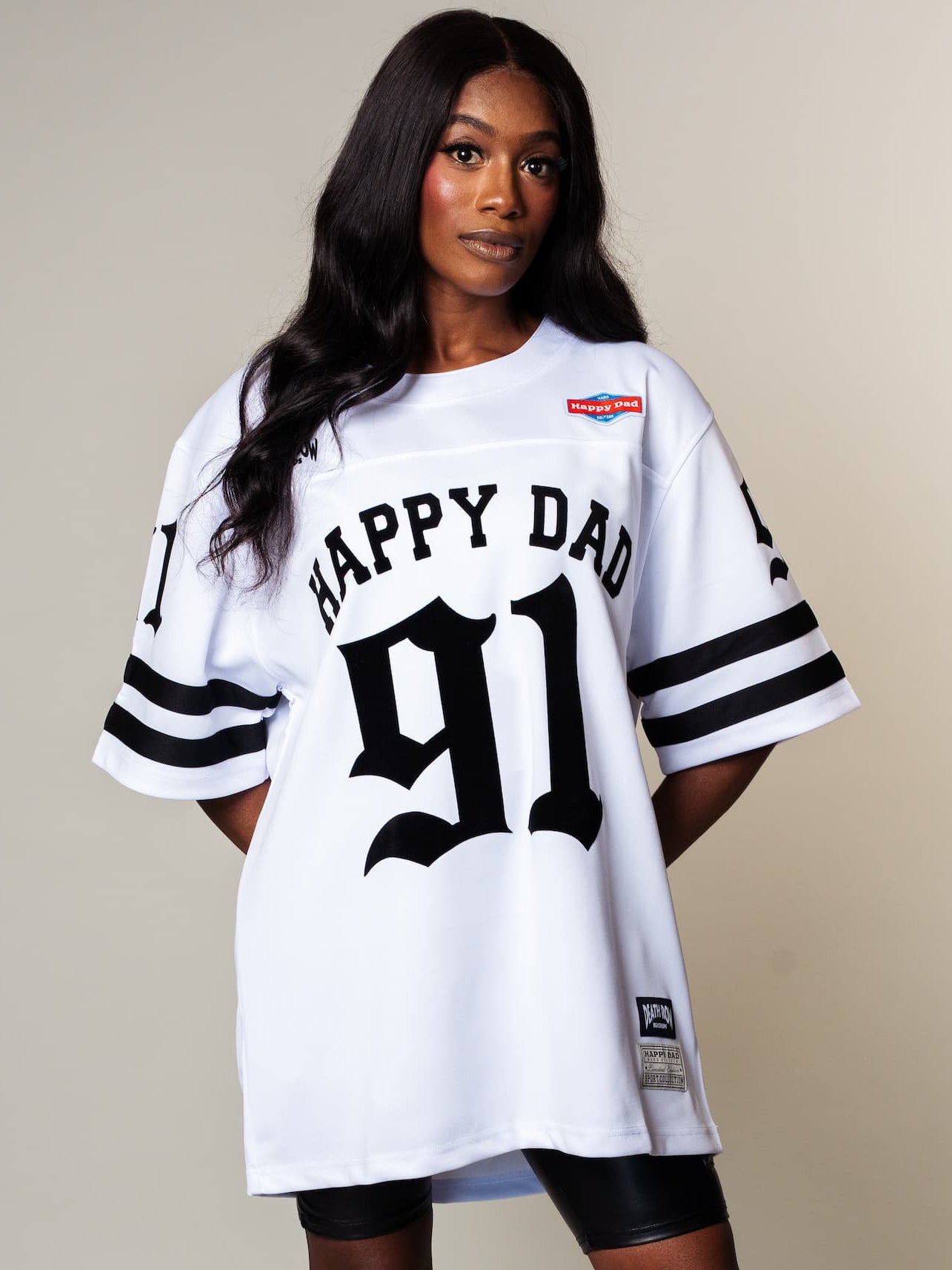 A woman models an oversized white sports jersey dress with 'HAPPY DAD 91' printed on the front, styled with black biker shorts and metallic brown knee-high boots against a plain beige studio background.