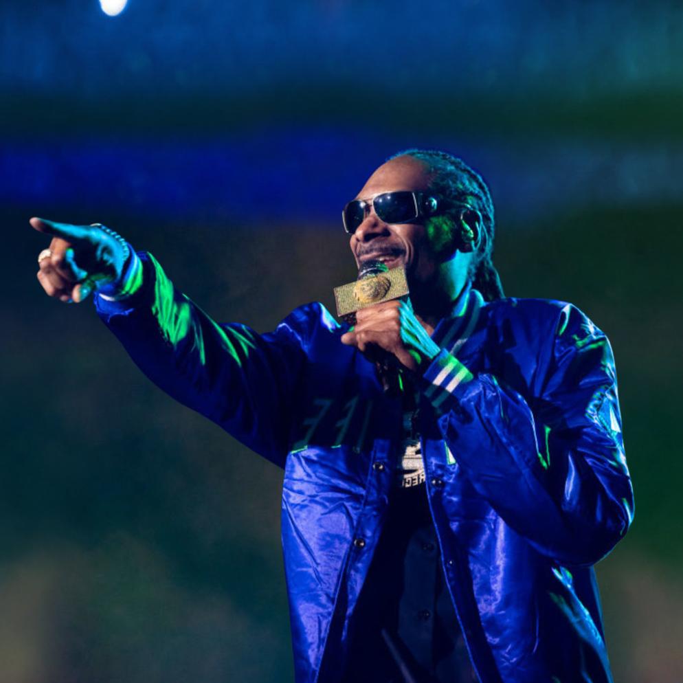 Snoop Dogg performing at a concert, wearing a jacket, holding the mic on one hand and pointing to the audience with the other hand while smiling