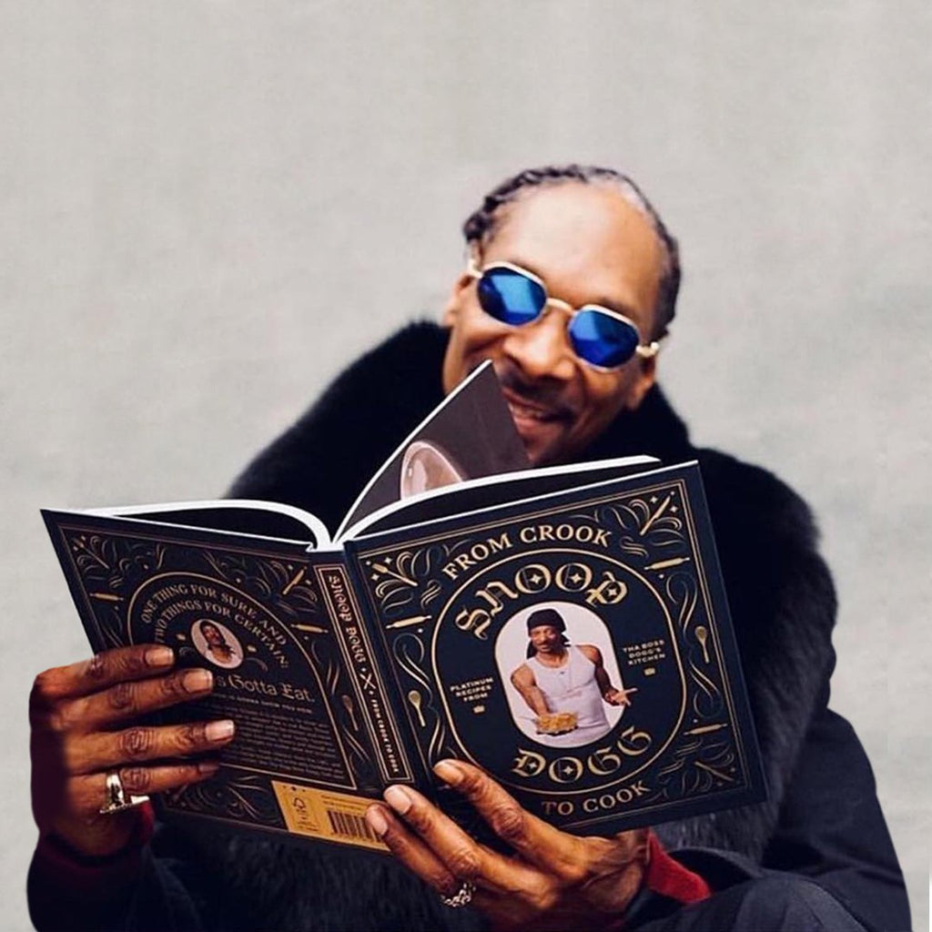 Snoop Dogg sitting down, reading his cooking book and smiling. He is wearing sun glasses